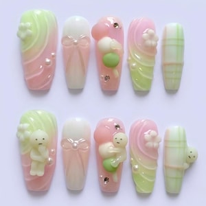 Op de afbeelding: Een set van tien kunstnagels in diverse ontwerpen. De nagels hebben pastelroze, groene en witte kleuren met decoratieve elementen zoals strikken, parels en kleine beeldjes. Sommige nagels hebben een gradi&euml;nteffect, andere een ruitpatroon.