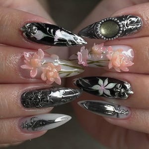 May include: A close-up of a set of long, pointed fingernails with intricate nail art. Designs include black and white floral patterns, 3D pink flowers, and silver metal embellishments. The nails showcase a variety of textures and styles.