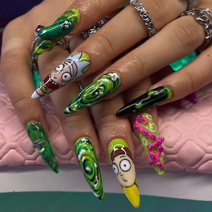 May include: Long, pointed acrylic nails with vibrant green, white, and black designs. Some nails feature cartoon character art, including a pickle and a character with yellow, white, and brown features. Other nails have swirling patterns and pink accents.