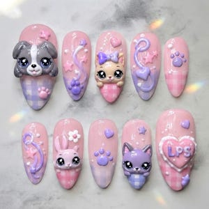 May include: A set of ten press-on nails featuring a pink and purple gingham pattern. The nails are decorated with 3D charms including animal faces, paw prints, stars, hearts, and flowers. One nail features a heart-shaped charm with the letters LPS.