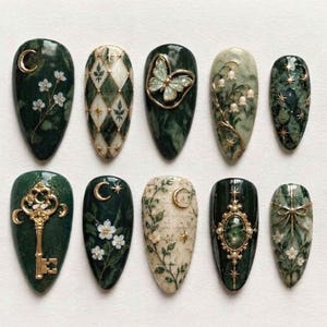 Op de afbeelding: Een set van tien amandelvormige press-on nagels met een kleurenpalet van donkergroen en crème. De ontwerpen bevatten gouden accenten, bloemmotieven, een vlinder, een vintage sleutel, maansikkels, sterren en gedetailleerde filigraanpatronen.