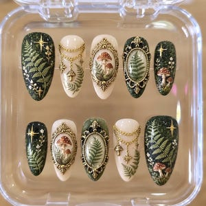 May include: A set of ten almond-shaped press-on nails in a clear plastic case. The designs feature dark green glitter backgrounds with fern and star motifs, cream-colored bases with gold chain charms, and ornate gold frames enclosing mushroom or fern art.