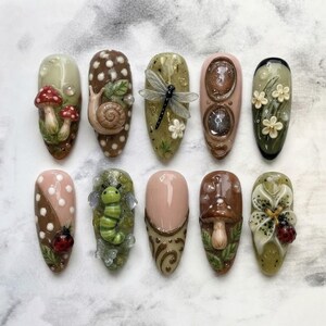 Pode incluir: Um conjunto de dez unhas postiças com designs em 3D, incluindo cogumelos, um caracol, uma libélula, flores, uma lagarta, uma joaninha e espirais. As unhas são decoradas com tons terrosos, incluindo nuances de verde, castanho e creme.