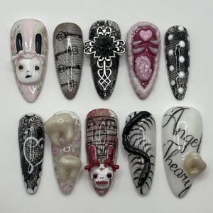 May include: A collection of ten long, oval-shaped artificial nails, each with a unique design. Designs include a bunny, keys, a cross, a heart, polka dots, teeth, musical notes, a snake, and the text "Angel Theory". The nails are painted in various colours, including pink, black, and white.