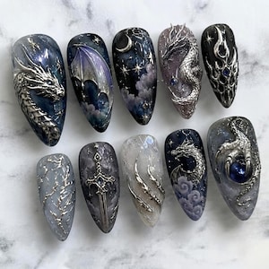 May include: A set of ten almond-shaped press-on nails featuring intricate 3D silver metallic designs. The motifs include detailed dragons, a sword, a crescent moon, stars, clouds, and a dragon claw clutching a dark blue sphere on a dark background.