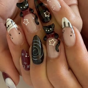May include: A set of almond-shaped, decorated fingernails. Designs include black cat figurines, star accents, and abstract patterns in black, white, and burgundy. Some nails feature a nude base with detailed art.