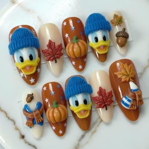 May include: A set of ten press-on nails featuring 3D autumn-themed designs. The nails include raised figures of Donald Duck wearing a blue knit hat, orange pumpkins, red and yellow maple leaves, brown acorns, and blue and white striped scarves.