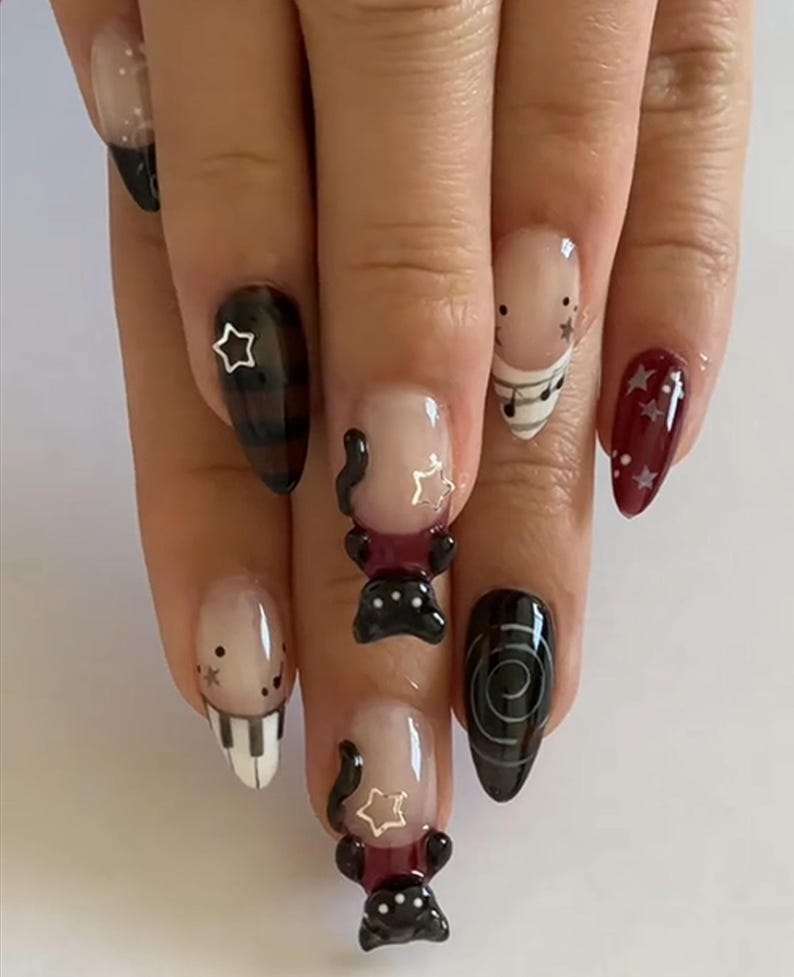 May include: A hand with a set of long, almond-shaped nails. The nails feature various designs, including a piano keyboard, musical notes, stars, and abstract black shapes. Some nails have a clear base with black tips.