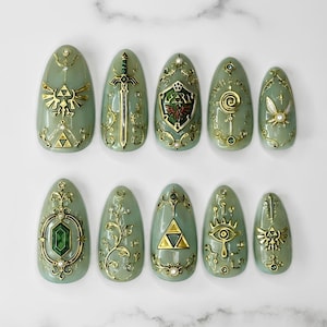 Peut inclure: Un ensemble de dix faux ongles en amande de couleur vert sauge, ornés de détails métalliques dorés complexes, notamment une épée, un bouclier, un symbole triforce, une fée, une pierre précieuse verte et un motif d'œil avec des perles.