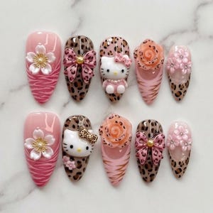 Peut inclure: Un ensemble de dix faux ongles en forme d'amande avec des motifs variés incluant des fleurs de cerisier roses, des imprimés léopard, des figurines Hello Kitty en relief, des roses pêche et des nœuds roses avec des perles dorées.