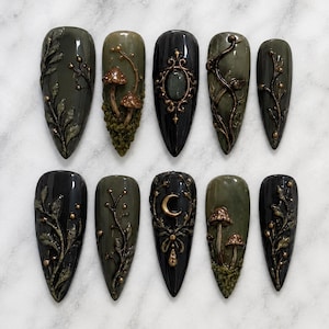 May include: A set of ten almond-shaped press-on nails in shades of olive green and black. The designs feature 3D embossed details including botanical leaf patterns, small brown mushrooms with textured moss, a central oval frame, and a gold crescent moon.