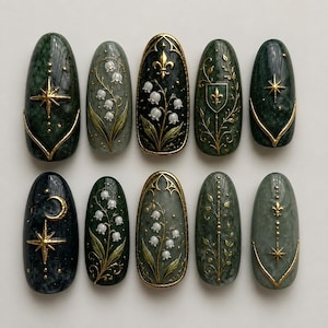 Op de afbeelding: Een set van tien kunstnagels met een donkergroene en zwarte marmerlook en gouden metalen accenten. De ontwerpen bevatten lelietjes-van-dalen, sterren, maansikkels, fleur-de-lis symbolen en verfijnde rankpatronen.