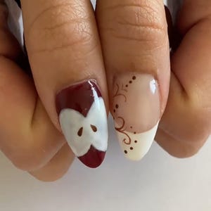 May include: Close-up of two fingernails with nail art. One nail features a red apple slice design with a white core and brown seeds. The other nail has a white tip with a brown swirl design and small dots.