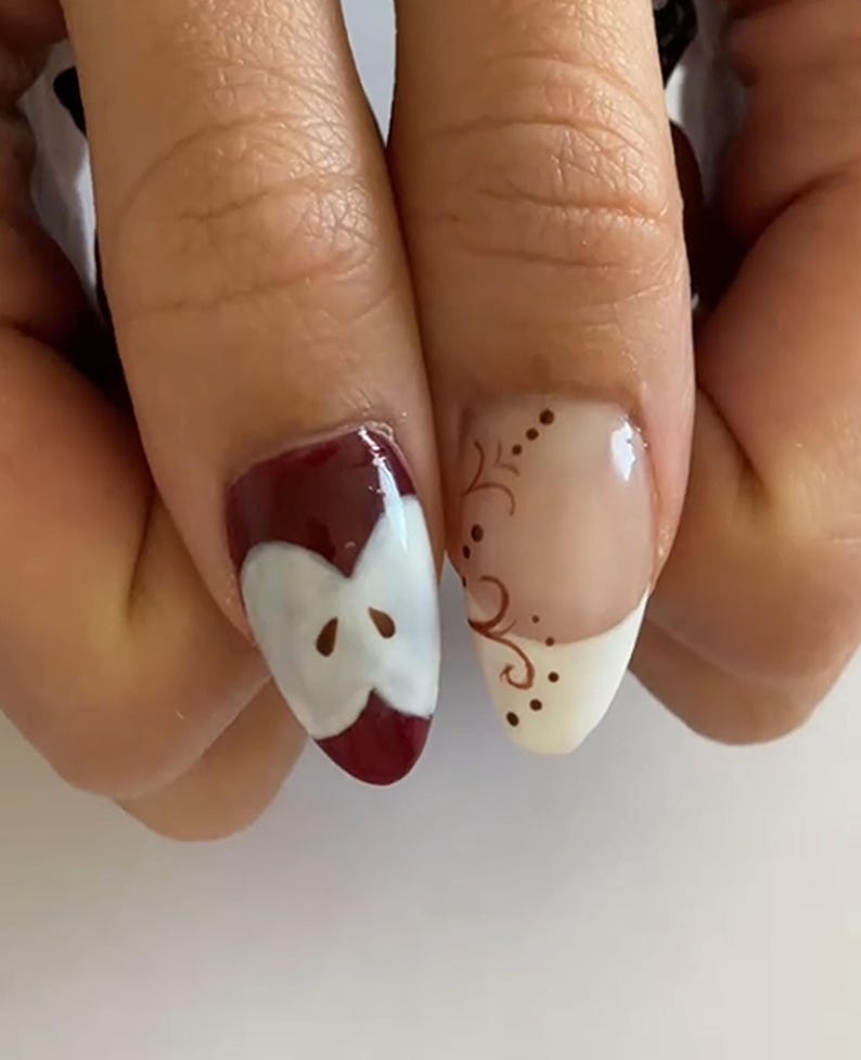 May include: Close-up of two fingernails with nail art. One nail features a red apple slice design with a white core and brown seeds. The other nail has a white tip with a brown swirl design and small dots.