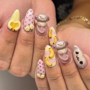 May include: A set of almond-shaped, decorated nails. The designs include 3D monkeys, bananas, and pink polka dots. The color palette features yellow, pink, and brown tones, creating a playful and whimsical aesthetic.