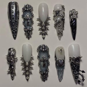 May include: A collection of ten long, pointed, and embellished artificial nails. Designs include silver spikes, crosses, skulls, and ornate detailing. The nails feature a mix of silver, black, and white colors, showcasing a gothic aesthetic.