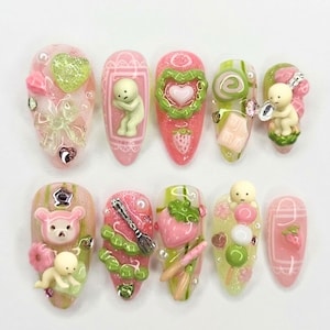 May include: A collection of ten artificial nails, each with a unique design. The nails are predominantly pink and green, adorned with 3D elements such as hearts, strawberries, and small figurines. Glitter and decorative patterns are also present.