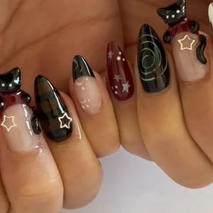 May include: A set of long, almond-shaped artificial nails. The nails feature a variety of designs, including black cat charms with star accents, a swirl design, and a burgundy nail with white stars. Some nails have a clear base with white accents.