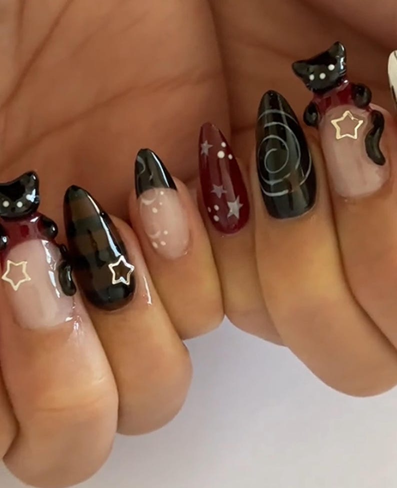 May include: A set of long, almond-shaped artificial nails. The nails feature a variety of designs, including black cat charms with star accents, a swirl design, and a burgundy nail with white stars. Some nails have a clear base with white accents.