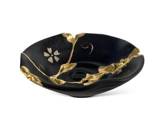 May include: A black ceramic bowl with gold detailing, demonstrating the Kintsugi technique. The bowl has a floral pattern and gold-filled cracks, emphasising its repaired state. The oval bowl shape and gold accents create a striking contrast. (approx. 20 cm)