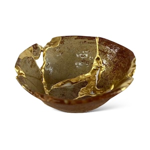 Può includere: Una ciotola in ceramica fatta a mano con una riparazione Kintsugi unica. La ciotola presenta un esterno marrone, un interno verde chiaro e accenti color oro che riempiono le crepe. La ciotola ha una forma organica e irregolare.
