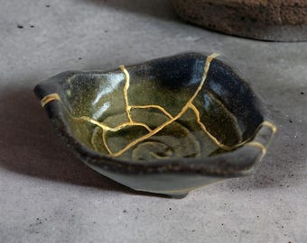 May include: A small, square ceramic bowl with a dark green and black glaze. The bowl features gold-coloured kintsugi, a Japanese art of repairing broken pottery with gold. The bowl is on a grey surface.