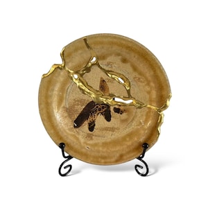 May include: A tan ceramic plate with a brown and gold design, repaired with gold-colored epoxy. The plate is displayed on a black metal stand, showcasing the Japanese art of Kintsugi.