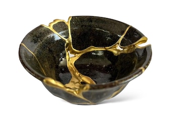 May include: A black ceramic bowl with gold-coloured detailing. The bowl's cracks are highlighted with gold, creating a striking contrast. The bowl has an irregular shape and a glossy finish, adding to its visual appeal.