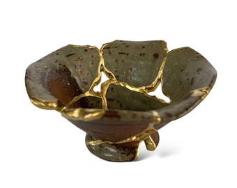 May include: A ceramic bowl with kintsugi repair. The bowl is brown and green with gold detailing. The bowl has a pedestal base and a petal-like shape, showcasing the Japanese art of repair.
