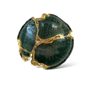 May include: A dark green ceramic bowl with gold-colored kintsugi repair. The bowl has a rounded shape with visible cracks filled with gold, creating a unique and artistic design. The bowl is set against a white background.