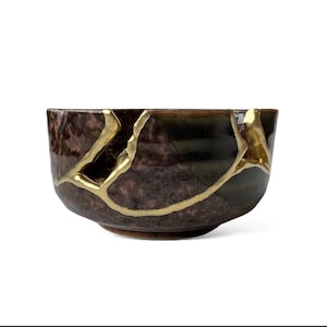 May include: A brown ceramic bowl with gold-colored kintsugi repair lines. The bowl has a dark, mottled finish with a rounded shape and a small foot. The gold lines highlight the cracks, creating a unique decorative piece.