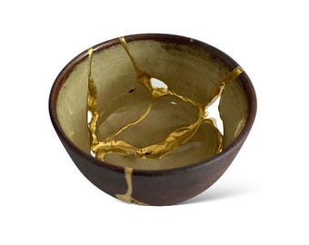 Peut inclure: Un petit bol rond en céramique, extérieur marron foncé et intérieur vert clair. Le bol est réparé avec du kintsugi doré, soulignant les fissures. Le bord est marron plus foncé, créant un contraste.