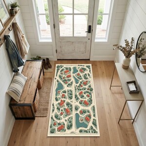 May include: A rectangular area rug featuring a top-down illustrated map of a neighborhood with winding roads, green trees, blue ponds, and small red-roofed houses. The rug is positioned on a light wood floor in an entryway with a wooden bench.