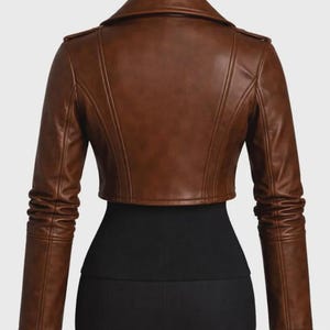 May include: A cropped brown jacket featuring a wide collar, long sleeves with gathered detailing at the forearms, and vertical seam lines across the back. The garment is displayed on a black mannequin torso against a plain background.
