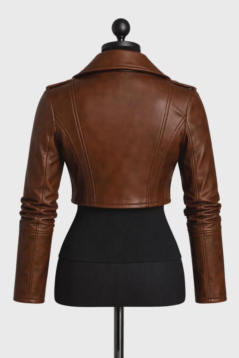 May include: A cropped brown jacket featuring a wide collar, long sleeves with gathered detailing at the forearms, and vertical seam lines across the back. The garment is displayed on a black mannequin torso against a plain background.