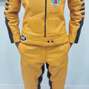 May include: Yellow leather jacket and pants set with black accents. The jacket has patches with text and logos, including "CAT Car Acrobatic Team" and "DM". The pants have ribbed detailing.