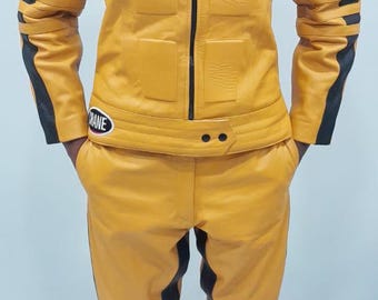 Può includere: Completo giacca e pantaloni in pelle gialla con dettagli neri. La giacca presenta toppe con testo e loghi, tra cui "CAT Car Acrobatic Team" e "DM". I pantaloni hanno dettagli a coste.