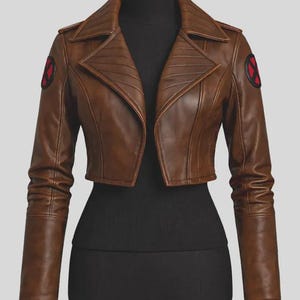 May include: A cropped brown jacket with long sleeves and a wide collar. The jacket features a circular black and red X-Men logo patch on each shoulder. The design includes structured paneling on the front and sleeves, with a smooth finish.