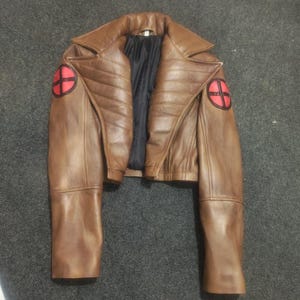 May include: A brown leather jacket featuring a wide collar, quilted detailing on the chest, and long sleeves. Each shoulder displays a circular red patch with a black cross design. The interior is lined with black fabric.