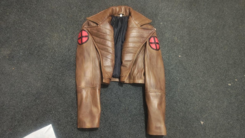 May include: A brown leather jacket featuring a wide collar, quilted detailing on the chest, and long sleeves. Each shoulder displays a circular red patch with a black cross design. The interior is lined with black fabric.
