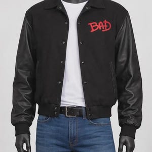 May include: Black varsity jacket with leather sleeves and the word "BAD" embroidered in red on the chest. The jacket has a button-up front, a black collar, and ribbed cuffs. The model is wearing blue jeans and a black belt.