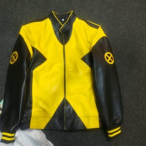 Pu&ograve; includere: Una giacca gialla e nera con un evidente design a forma di X sul davanti. Le maniche sono nere con polsini a righe gialle e un logo circolare giallo con una X su ogni spalla. La giacca presenta un colletto alto e una chiusura a zip.