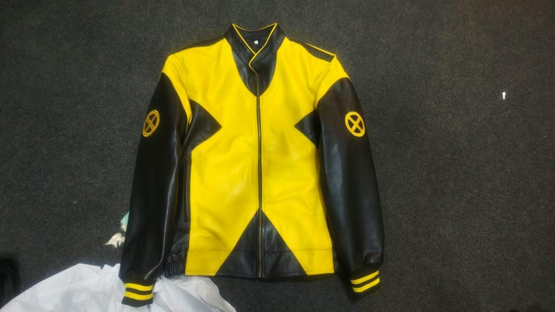 Pu&ograve; includere: Una giacca gialla e nera con un evidente design a forma di X sul davanti. Le maniche sono nere con polsini a righe gialle e un logo circolare giallo con una X su ogni spalla. La giacca presenta un colletto alto e una chiusura a zip.