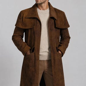 May include: A long brown suede coat featuring a wide collar, a cape-style overlay across the shoulders, and two deep front pockets. The coat is worn over a beige crew-neck sweater and tan trousers, creating a layered look with a textured finish.