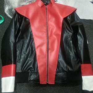 May include: A black faux leather jacket featuring a prominent red V-shaped panel extending from the collar to the hem. The sleeves include red and white color-blocked cuffs, a full-length front zipper, and a black ribbed waistband.