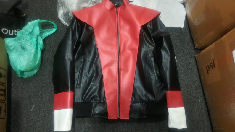 May include: A black faux leather jacket featuring a prominent red V-shaped panel extending from the collar to the hem. The sleeves include red and white color-blocked cuffs, a full-length front zipper, and a black ribbed waistband.