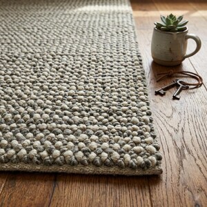 May include: A textured, neutral-toned area rug with a speckled pattern. The rug features small, raised bumps in shades of grey and off-white. A small succulent plant in a ceramic mug and a pair of antique keys sit on the wooden floor beside the rug.