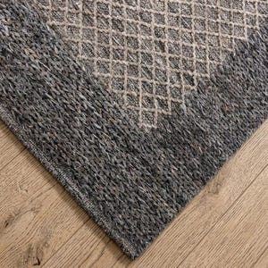 Puede incluir: Una esquina de una alfombra tejida gris y beige con un patrón de diamantes. La alfombra tiene una apariencia texturizada y está colocada sobre un suelo de madera marrón claro.