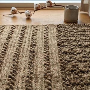 May include: A brown rectangular rug with a textured surface. The rug has a combination of vertical striped and textured patterns. The rug is made of a natural fibre and is placed on a wooden floor. A small vase with cotton stems is in the background.