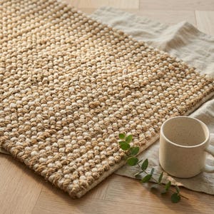 Puede incluir: Una alfombra texturizada de color beige y crema con un diseño tejido con nudos. Una taza de color crema con un acabado moteado se encuentra sobre un paño de lino junto a la alfombra. Una ramita de hojas verdes añade un toque natural.